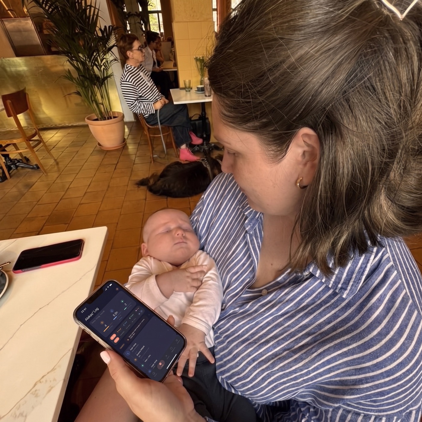 A parent holding a sleeping newborn in a caf&eacute;, checking the day's log on her phone.